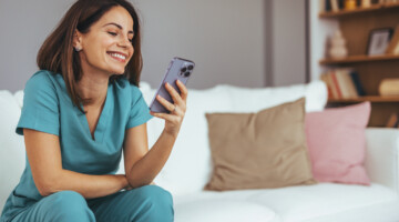 A cheerful woman in medical scrubs sits on a sofa, engaging with her smartphone. The cozy living room setting adds warmth and comfort to the scene, highlighting relaxation and connectivity.