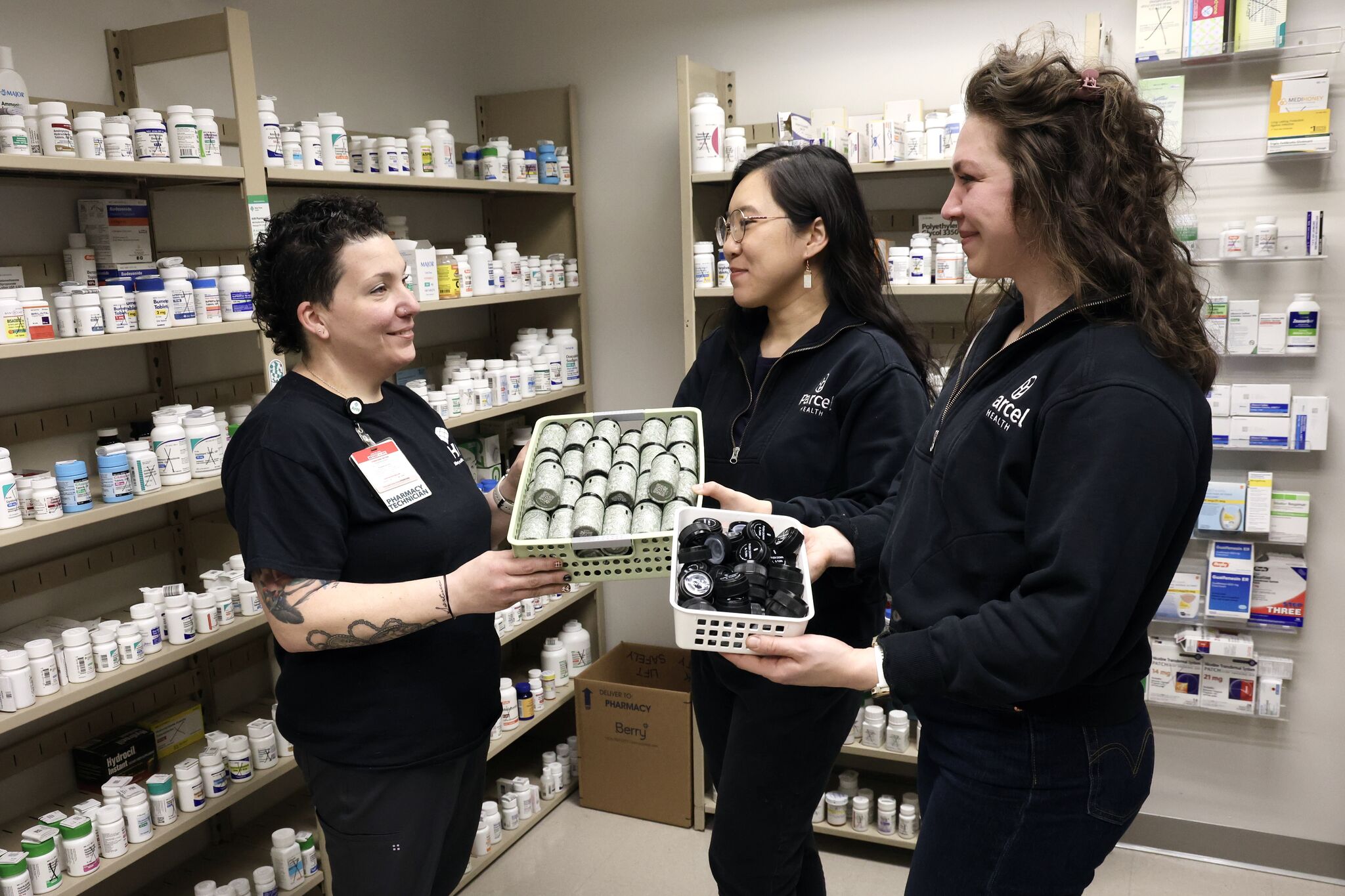 Three people in a pharmacy discussing reusable pill containers while standing among shelves of medication bottles.